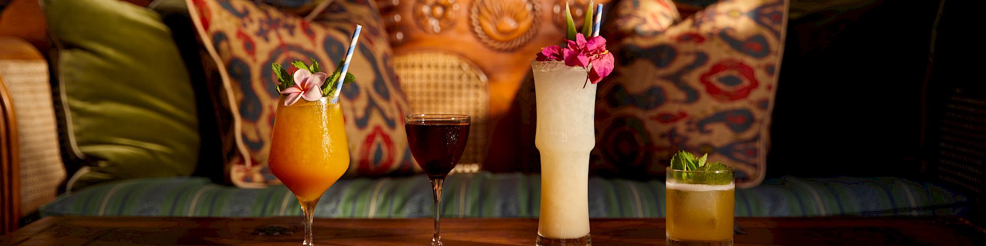 The image shows a cozy seating area with colorful cushions and a wooden bench, featuring four different types of drinks on a table, each garnished uniquely.