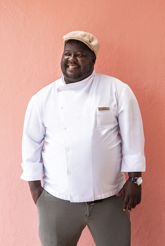 A person in a chef's uniform and cap is standing against a pink wall, smiling with hands in pockets.