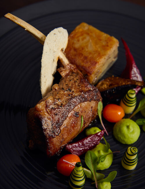 A gourmet dish with lamb chops, layered pastry, colorful vegetables, and artful garnishes, presented on a black plate.