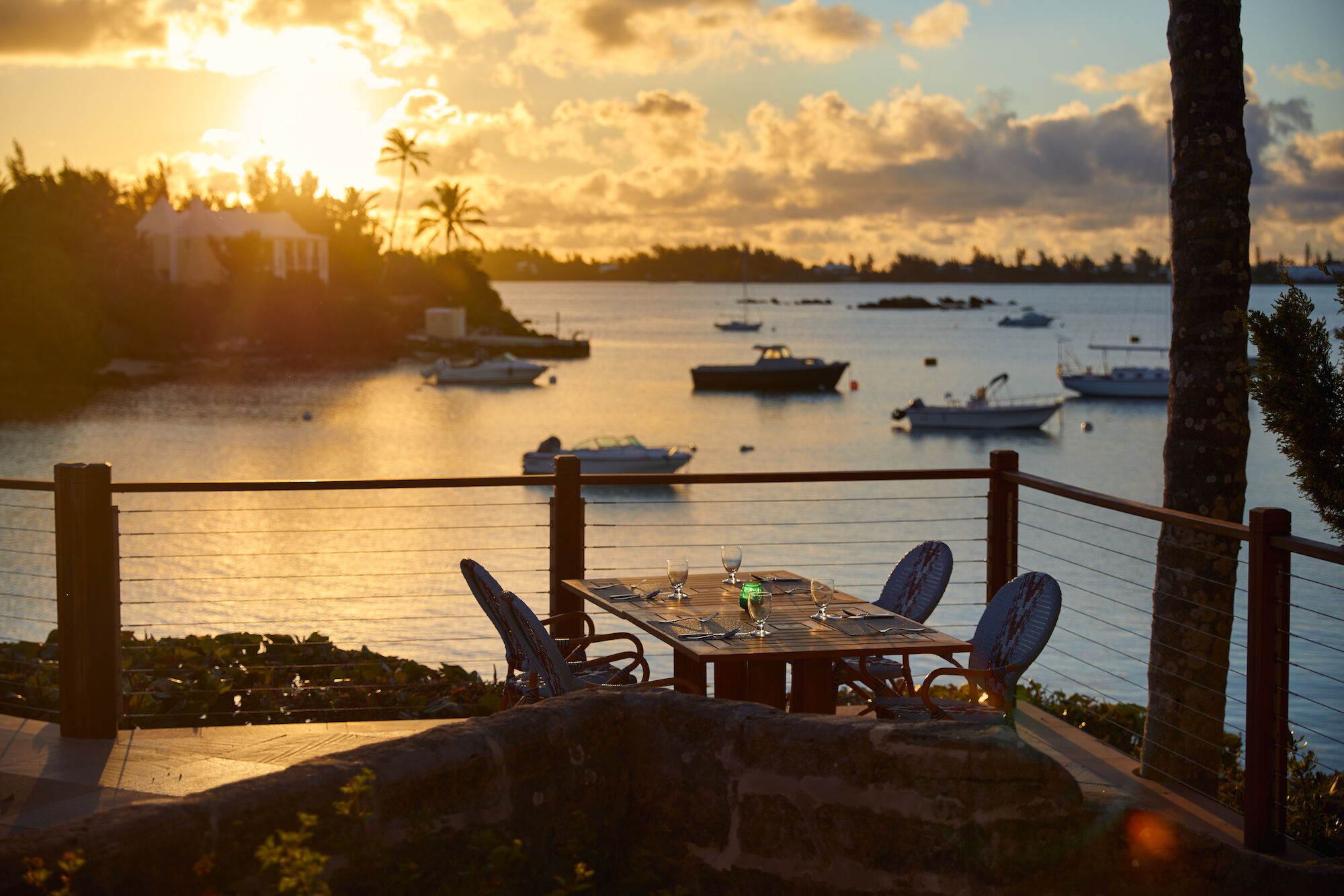 A table with glasses sits on a patio overlooking a serene bay with boats and a sunset in the background, surrounded by tropical trees.