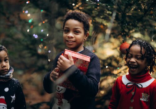 Three children are standing in front of a decorated Christmas tree. One child is holding a wrapped gift, and festive lights are visible.