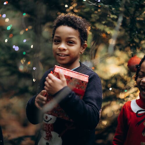 Three children are standing in front of a decorated Christmas tree. One child is holding a wrapped gift, and festive lights are visible.