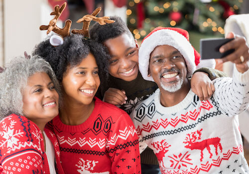 A family wearing festive sweaters and accessories takes a selfie in front of a decorated Christmas tree, smiling and celebrating the holiday.