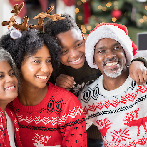 A family wearing festive sweaters and accessories takes a selfie in front of a decorated Christmas tree, smiling and celebrating the holiday.