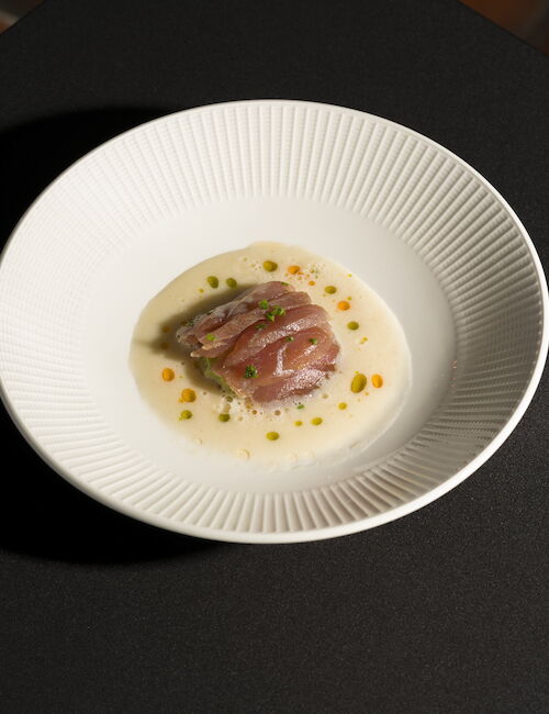 The image shows a dish of sliced fish on a white plate, garnished with sauce and small colorful dots, placed on a dark surface.