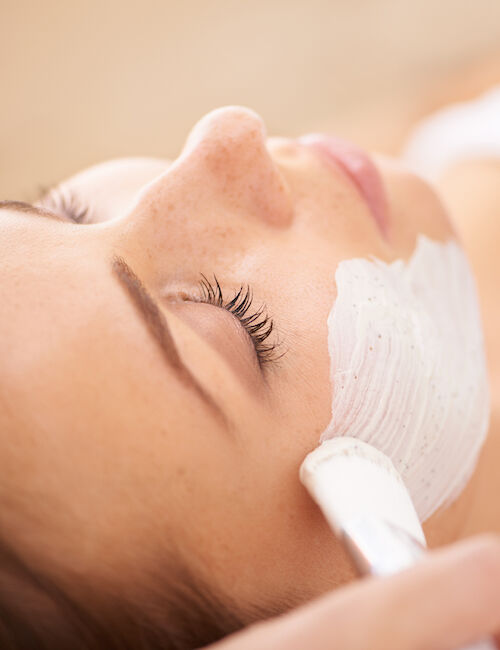 A person receiving a facial treatment, with a white cream being applied to their face using a brush, is visible in the image.