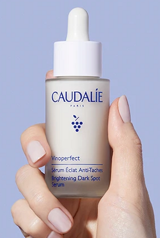 A hand is holding a bottle of Caudalie Vinoperfect Brightening Dark Spot Serum against a light blue background.