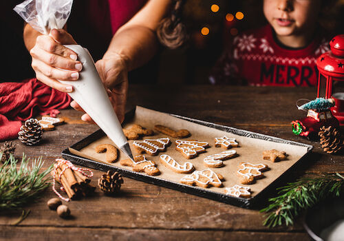 A person is decorating Christmas-themed cookies with icing on a tray, surrounded by holiday decorations like pine cones and greenery.