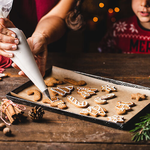 A person is decorating Christmas-themed cookies with icing on a tray, surrounded by holiday decorations like pine cones and greenery.