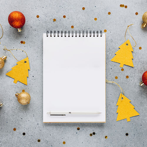 A spiral notebook and pen are surrounded by gold and red Christmas ornaments and tree-shaped tags on a gray surface, with scattered confetti.