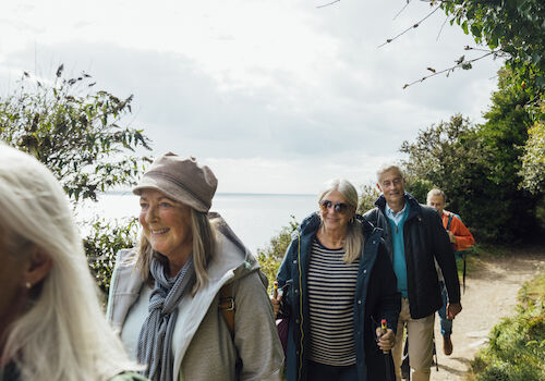 A group of smiling people hiking on a scenic, tree-lined path by a body of water under a cloudy sky.