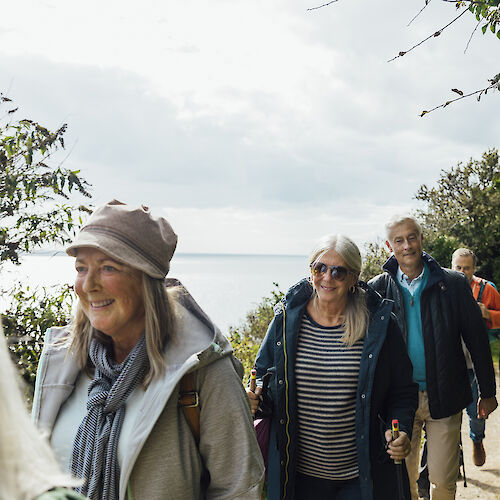 A group of smiling people hiking on a scenic, tree-lined path by a body of water under a cloudy sky.