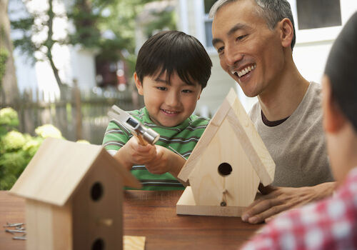 A child and an adult are building wooden birdhouses together at a table outside, enjoying a hands-on activity in a garden setting.