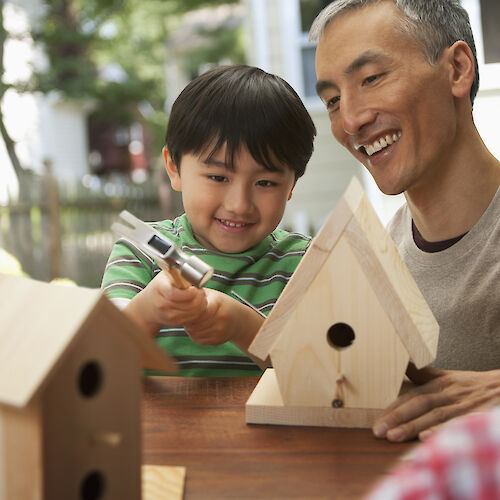 A child and an adult are building wooden birdhouses together at a table outside, enjoying a hands-on activity in a garden setting.