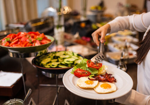 A person serves food onto a plate at a buffet, adding tomatoes and greens beside sunny-side-up eggs and bacon.