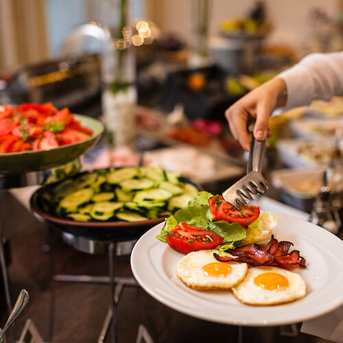 A person serves food onto a plate at a buffet, adding tomatoes and greens beside sunny-side-up eggs and bacon.