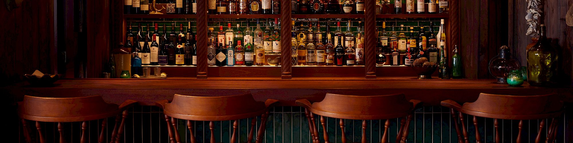 The image shows a dimly-lit bar with wooden chairs and shelves filled with various liquor bottles, creating a cozy and inviting atmosphere.