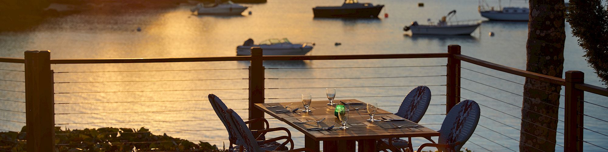 A scenic waterfront view at sunset with a dining table set up on a patio, overlooking boats on the water and surrounded by palm trees and foliage.