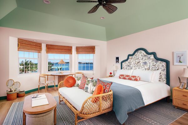 A cozy bedroom with a large bed, wicker furniture, and ocean view through windows. Light decor with pastel colors and a ceiling fan.