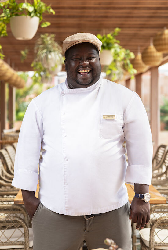 A chef in a white coat and cap stands in a decorated outdoor dining area with wicker chairs and hanging plants, smiling at the camera.