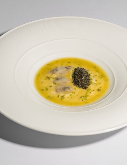 A white plate with a gourmet dish, featuring ravioli in a yellow sauce and topped with a small mound of caviar, garnished with herbs.