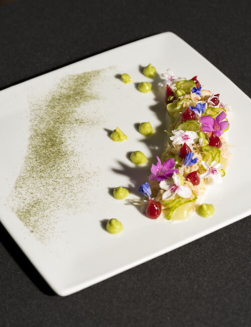 A gourmet dish on a white square plate, featuring colorful edible flowers, avocado dots, and a sprinkle of green powder.