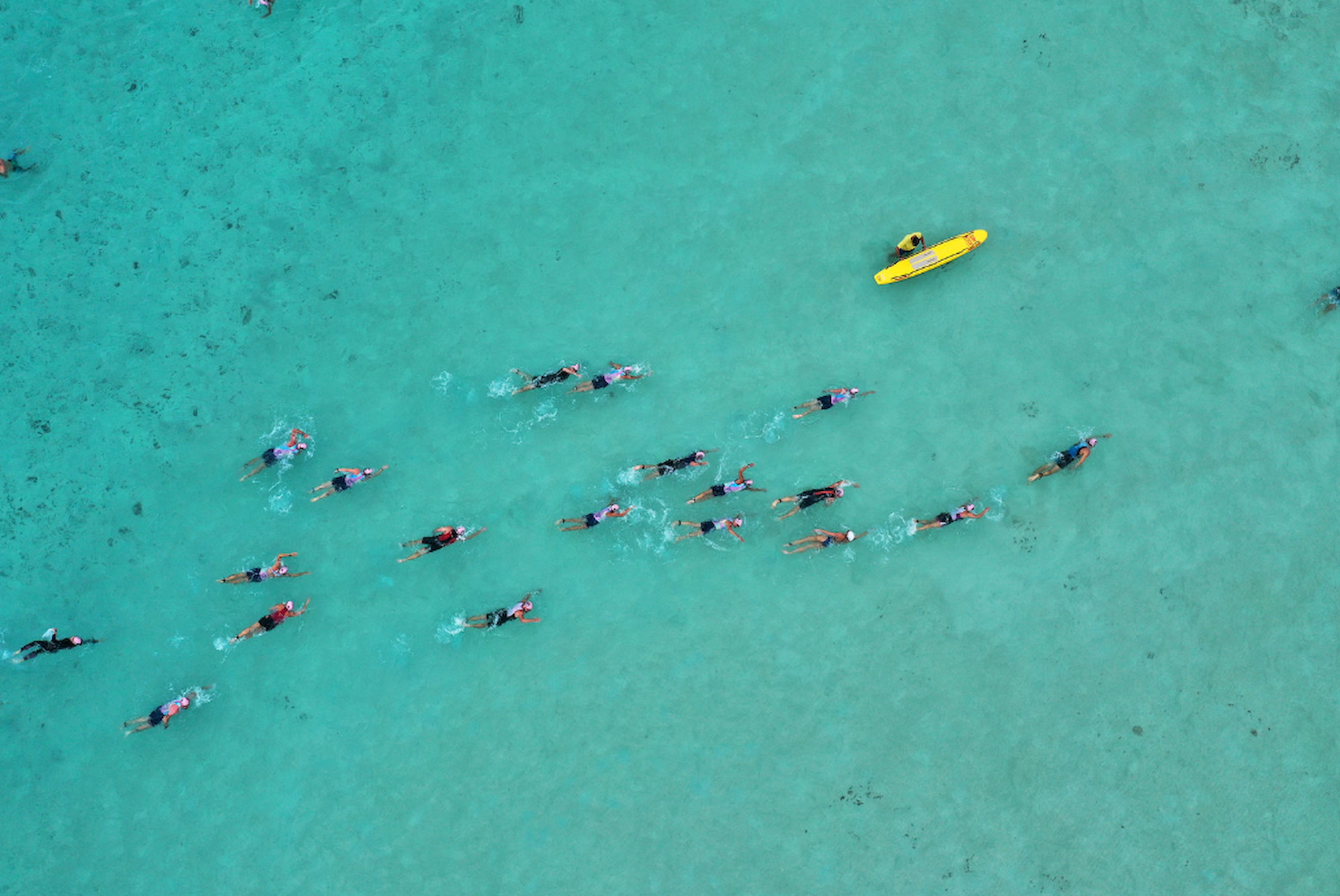 People swimming in clear turquoise water alongside a yellow kayak, viewed from above.