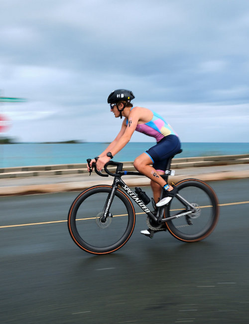 A cyclist in motion on a road by the sea, wearing a helmet and colorful attire, with a blurred background showing signs and a cloudy sky.