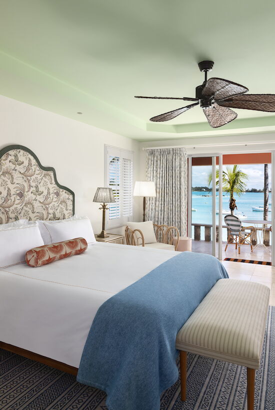 A cozy bedroom with a coastal view features a bed, armchairs, ceiling fan, and desk, opening to a balcony overlooking the ocean.