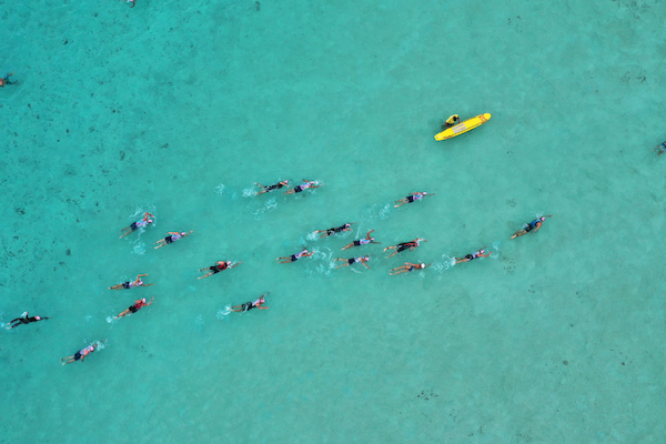 Aerial view of swimmers in clear turquoise water, accompanied by a yellow kayak.