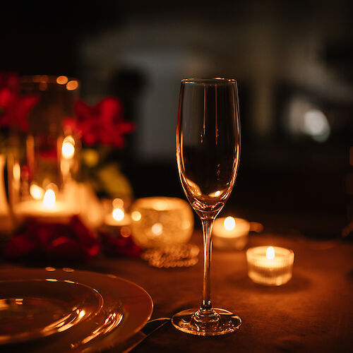 A dimly lit table with a champagne glass, plates, and candles, surrounded by small floral decorations, creating a romantic ambiance.