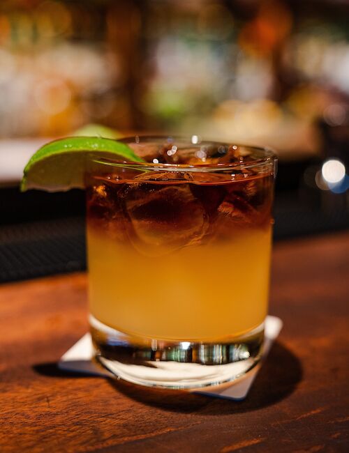 A cocktail with a lime wedge garnish sits on a wooden bar counter, featuring a dark layered top over a lighter liquid base.