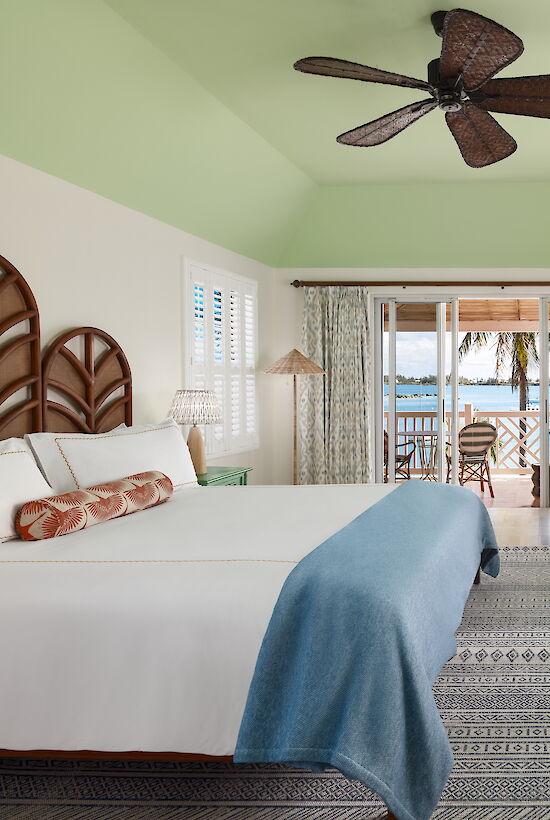 A bright bedroom with a bed, sofa, ceiling fan, and a view of a balcony leading to a palm tree-lined waterfront.