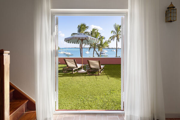 A view from inside a house shows a patio with two lounge chairs, a sun umbrella, grass, palm trees, and a glimpse of the ocean beyond.