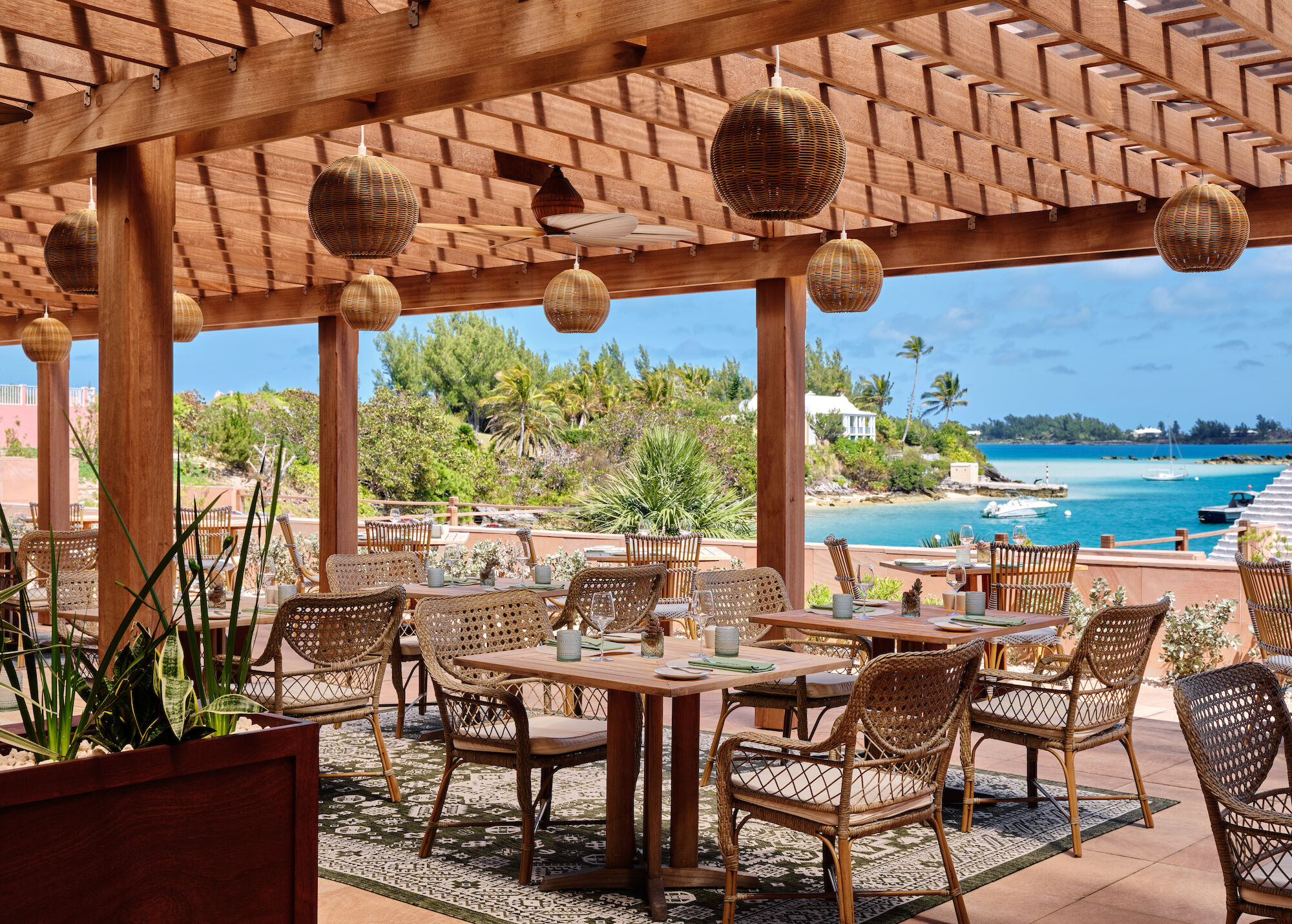 An outdoor dining area with wicker chairs under a pergola, tropical plants, and a view of the ocean and distant greenery completes the scene.