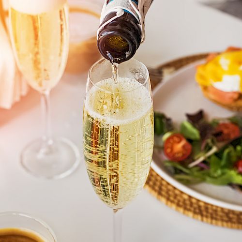 A bottle is pouring sparkling wine into a glass. There are also other glasses and a plate with a salad and an egg dish in the background.