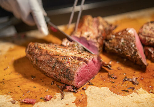 A hand slices a seasoned, cooked piece of meat on a wooden board, showing juicy, tender pink insides in a kitchen setting.