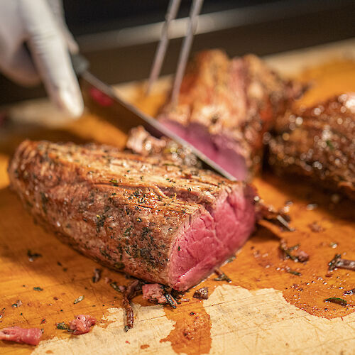 A hand slices a seasoned, cooked piece of meat on a wooden board, showing juicy, tender pink insides in a kitchen setting.