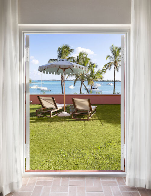 A view through an open doorway shows a grassy area with two lounge chairs and an umbrella, overlooking the ocean with palm trees.