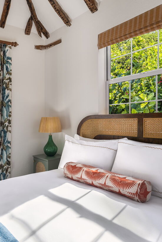 A cozy bedroom features a bed, colorful curtains, a side table with a lamp, and double doors opening to a sunny brick patio.