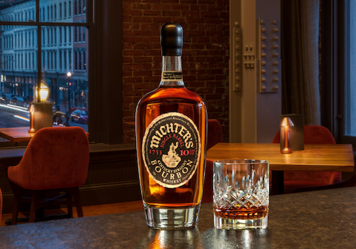 A bottle of Michter's bourbon and a glass are displayed on a table in a cozy bar setting, with warm lighting and city views.