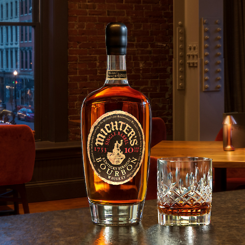 A bottle of Michter's bourbon and a glass are displayed on a table in a cozy bar setting, with warm lighting and city views.