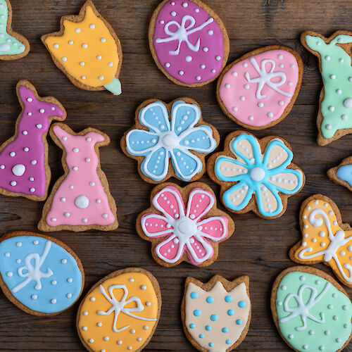 Colorful, decorated cookies shaped like butterflies, flowers, dresses, and eggs are arranged on a wooden surface.