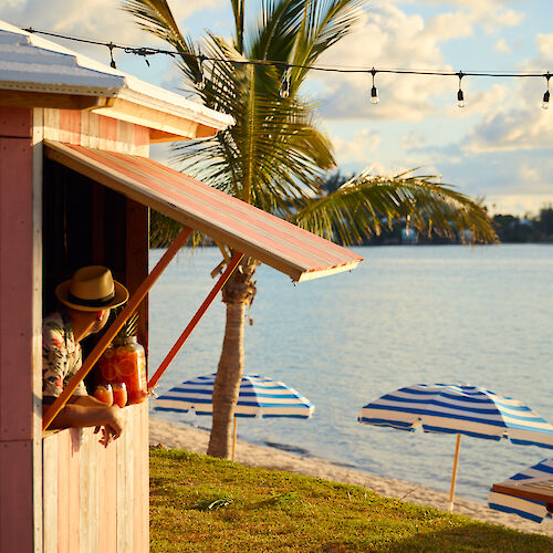 A small pink beach shack with a person inside, palm trees, string lights, and striped umbrellas by the water under a blue sky.