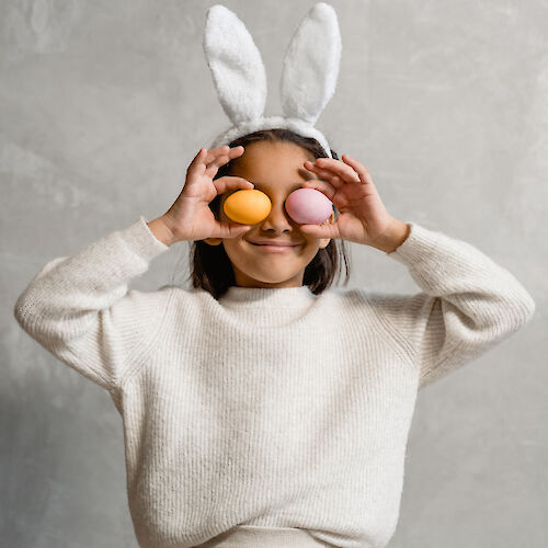 A child wearing bunny ears holds colored eggs in front of their eyes, smiling playfully against a neutral background.