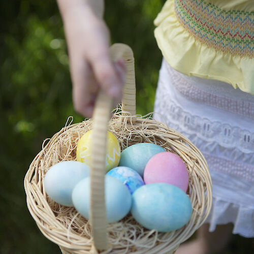 A person is holding a basket filled with pastel-colored Easter eggs, standing on grass.