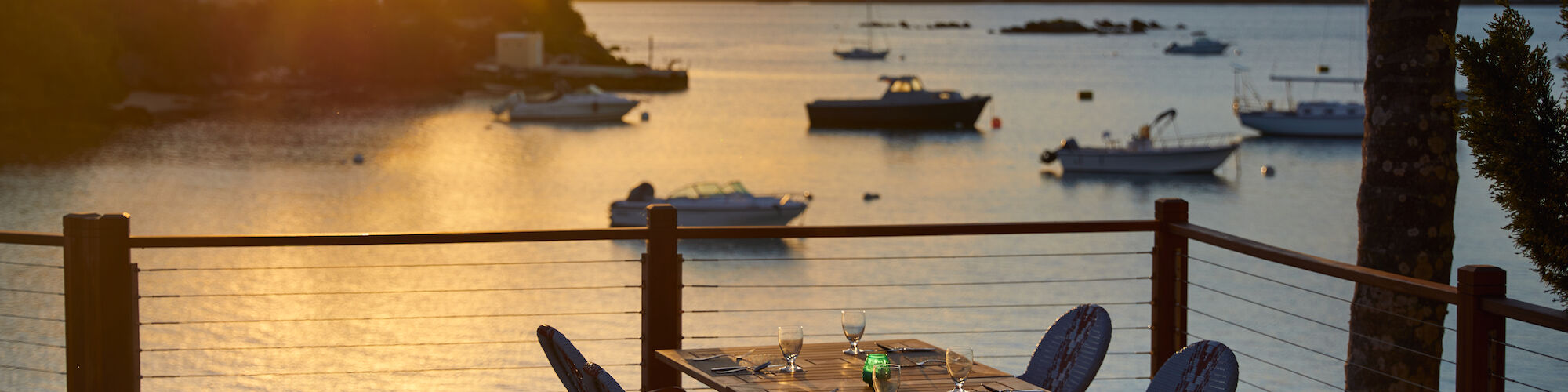 A scenic sunset view over a calm body of water with boats. In the foreground, there's an outdoor dining table set with chairs.