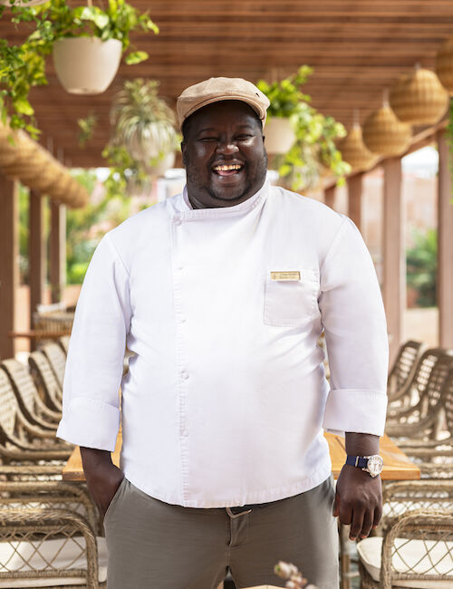 A chef in a white coat and cap stands in a decorated outdoor dining area with wicker chairs and hanging plants, smiling at the camera.