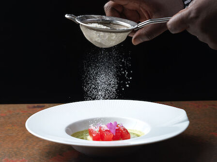 A person is dusting powdered sugar over a dessert with flowers and fruit in a white plate, using a fine mesh sieve.