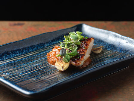 The image shows a piece of grilled fish garnished with herbs and vegetables on a rectangular blue plate.